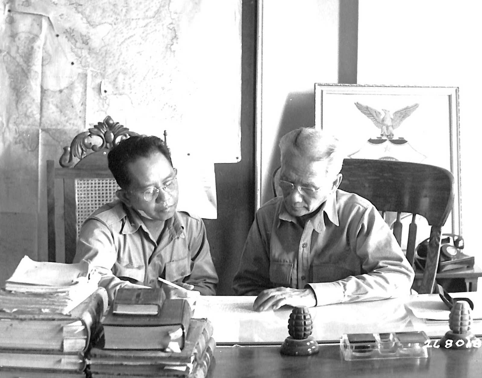 Photo of Carlos Romulo and  Sergio Osmeña sitting at a table covered in books, reading a document